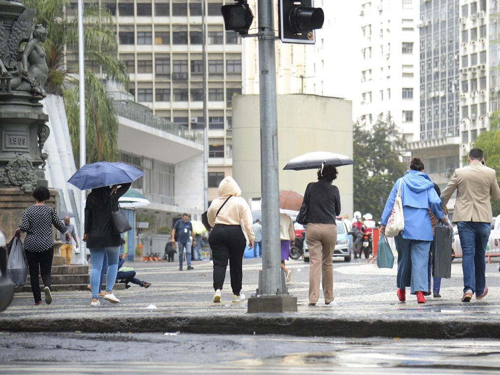 Pessoas agasalhadas caminhando em rua de São Paulo durante dia de céu nublado e temperaturas amenas.