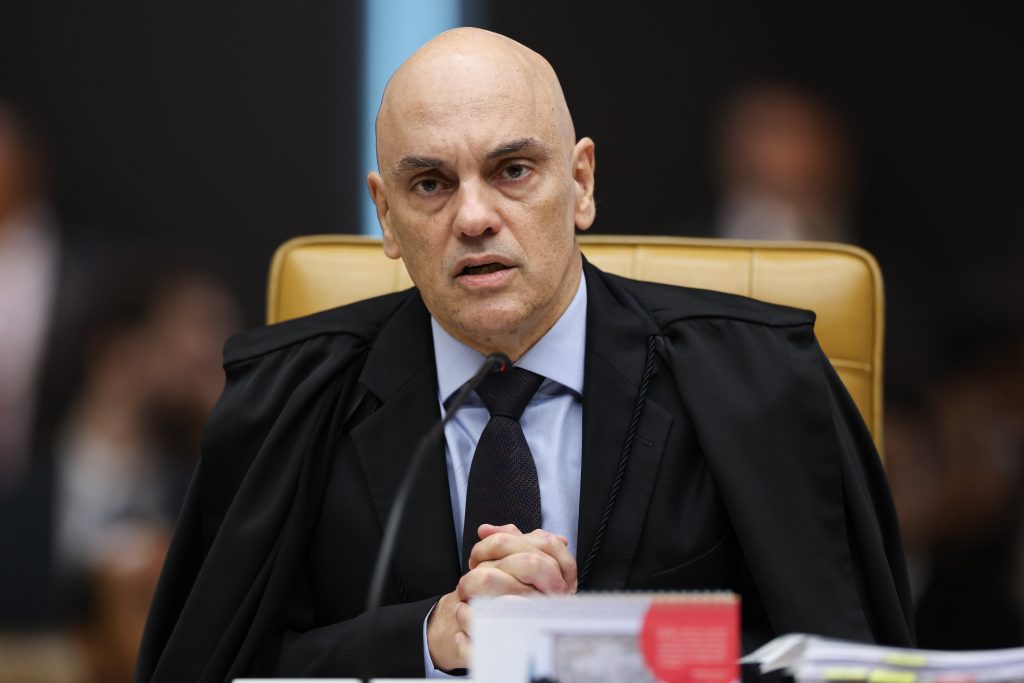 Alexandre de Moraes (STF) em sessão no plenário, usando toga preta e camisa social, sentado à mesa com microfone e expressão séria. STF visto.
