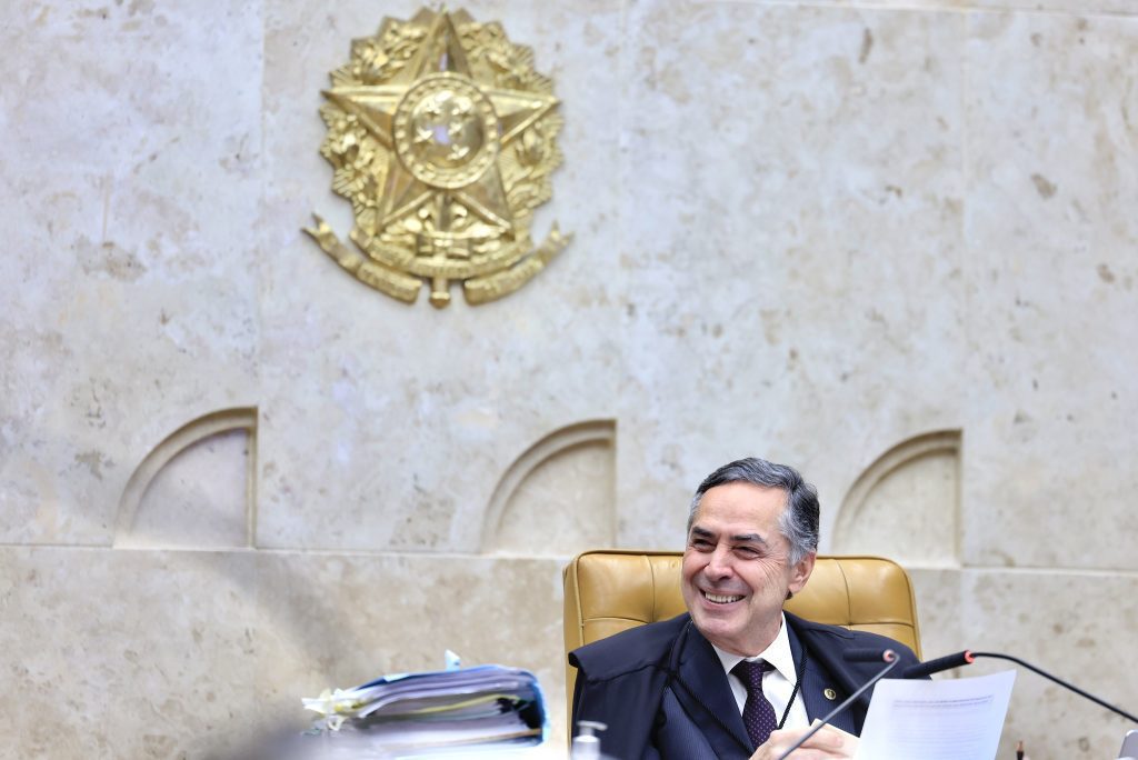 STF visto: Ministro Luís Roberto Barroso, atual Presidente do STF, sentado em sessão no plenário com mesa de trabalho e microfone, ao lado do brasão do Supremo Tribunal Federal no fundo.