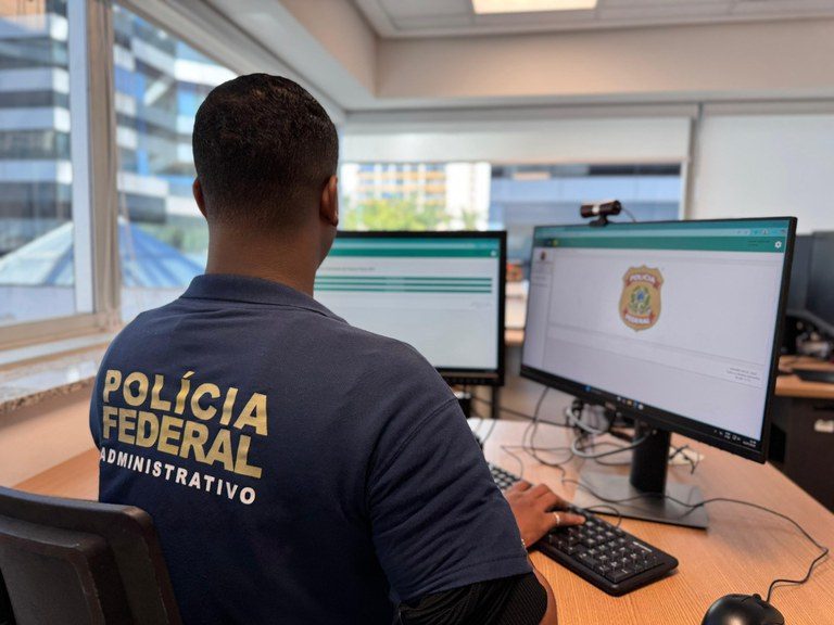 Agente da Polícia Federal Administrativa em uma sala com computadores, monitorando sistemas, com texto “POLÍCIA FEDERAL” nas costas; MPF aciona União por ausência de comprovação na transferência do controle de CACS à Polícia Federal.