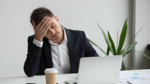 Homem em escritório com expressão de cansaço e estresse, segurando o rosto e olhando para o computador, enquanto uma xícara de café está na mesa; imagem sobre Burnout no Brasil em contexto de trabalho no varejo, educação e marketing.