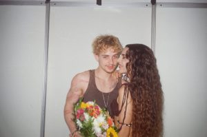 Juliano Floss e Marina Sena em foto romântica durante evento, com ele segurando um buquê colorido e ela de frente, em ambiente interno.