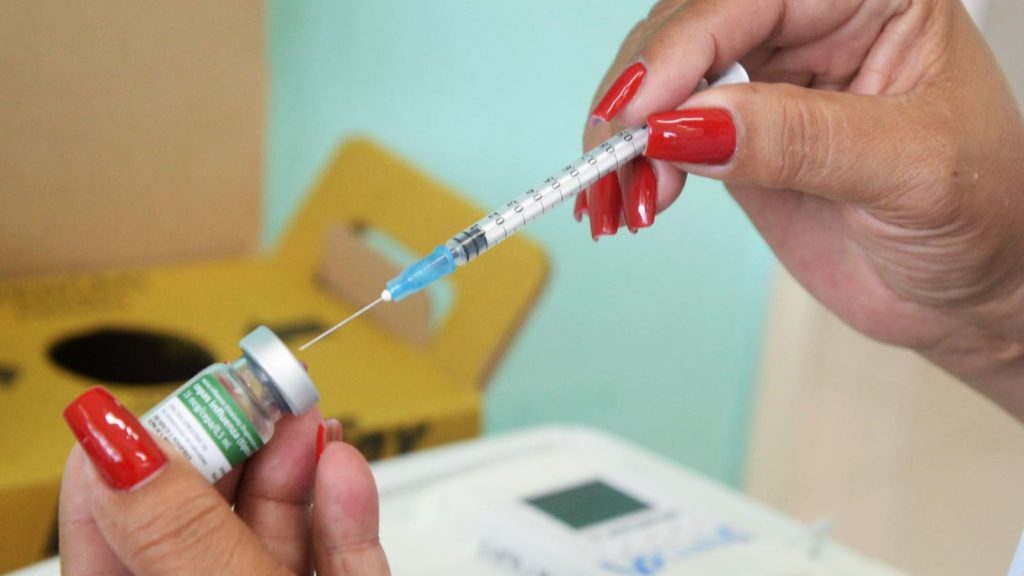 Vacinação neste fim de semana: profissional segura seringa e frasco de vacina, com agulha pronta para aplicação, em atendimento de saúde.