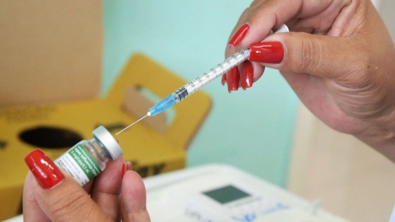 Vacinação neste fim de semana: profissional segura seringa e frasco de vacina, com agulha pronta para aplicação, em atendimento de saúde.