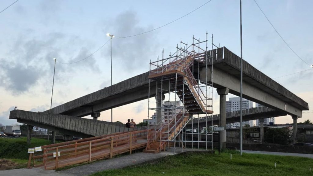 Obras em viaduto com estrutura metálica e andaimes na pista, ao lado de vias sob céu nublado. Pacote CNL anuncia investimento nas rodovias da Baixada Santista e Vale do Ribeira com 90 km de vias marginais na SP-055.