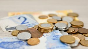 Moedas e cédulas brasileiras sobre um fundo desfocado, representando o mercado e a Projeção da inflação para 2026 com redução para 5,15%.