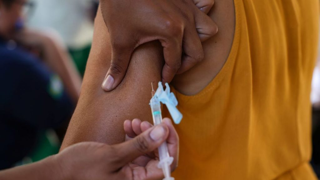 Vacina sendo aplicada em braço de uma pessoa, com seringa e profissional segurando o aplicador; contexto de prevenção ao sarampo, com foco em casos em São Paulo e maioria de bebês.