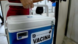 Caixa térmica de vacina com o texto “VACINA”, em um ambiente de campanha de saúde, destacando a necessidade de prevenção e atenção às mortes por gripe em Campinas