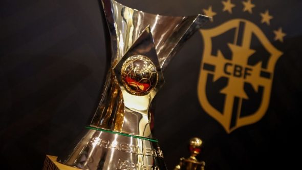 Taça de premiação dourada ao lado do escudo da CBF em um cenário esportivo, sugerindo a pergunta “Brasileirão volta quando?” com datas e retomada após eliminação do Brasil.