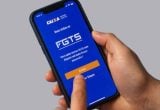 FGTS em um celular com o aplicativo Caixa aberto, com um dedo tocando a tela; a imagem destaca a mensagem sobre distribuição de R$ 13 bilhões e a possibilidade de recebimento.