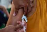 Vacina sendo aplicada em braço de uma pessoa, com seringa e profissional segurando o aplicador; contexto de prevenção ao sarampo, com foco em casos em São Paulo e maioria de bebês.