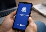 Pessoa segura um celular com a tela exibindo a Carteira de Trabalho Digital e a mensagem sobre desemprego, indicando queda e menor nível da série para o período.