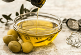 Azeite de oliva sendo derramado em uma tigela de vidro, com azeitonas ao lado, remetendo a alertas e testes contra fraudes em azeites de oliva.