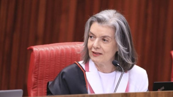 Cármen Lúcia em sessão solene, sentada em cadeira de couro vermelho, diante de microfone, com expressão serena, em ambiente institucional.