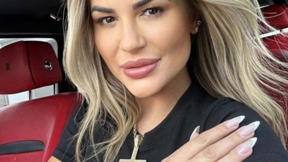 Deolane Bezerra em foto de close, com cabelo loiro e maquiagem marcada, usando camiseta preta com pingente cruciforme, sentada em veículo com bancos vermelhos ao fundo. OAB-SP suspendeu Deolane Bezerra e o julgamento deve ocorrer em até 360 dias.