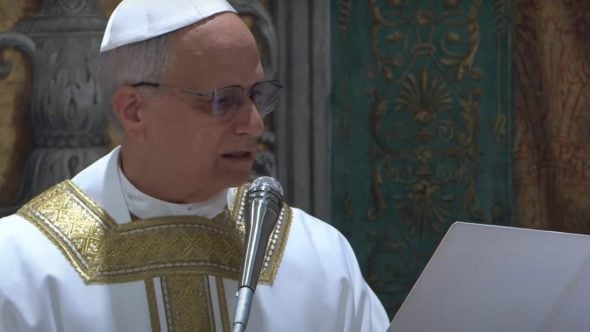 Papa Leão 14 em cerimônia religiosa, usando paramentos dourados e mitra, diante de um microfone e de um documento, em ambiente interno.