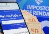 Quarto lote imposto de renda: Pessoa consultando site da Receita Federal no celular com cartelas de dinheiro de 50 e 100 reais ao fundo; tema do Imposto de Renda, com informação sobre o Quarto lote da restituição do Imposto de Renda.