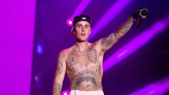 Justin Bieber aparece em palco, com tatuagens aparentes, boné e correntes, em um show com luzes roxas e feixe de holofotes, em contexto de confirmação na final da Copa do Mundo.