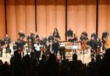 Orquestra Sinfônica de Campinas em ensaio ao lado de regente e músicos, com violinos e instrumentos de sopro em palco iluminado, apresentando música para o público.