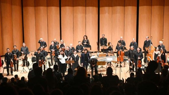 Orquestra Sinfônica de Campinas em ensaio ao lado de regente e músicos, com violinos e instrumentos de sopro em palco iluminado, apresentando música para o público.