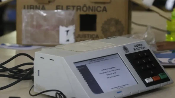 Urna eletrônica em preparação para votação, com teclado numérico e visor na tela, ao lado de equipamentos de apoio. Tema: Eleições 2026 e segurança auditável.
